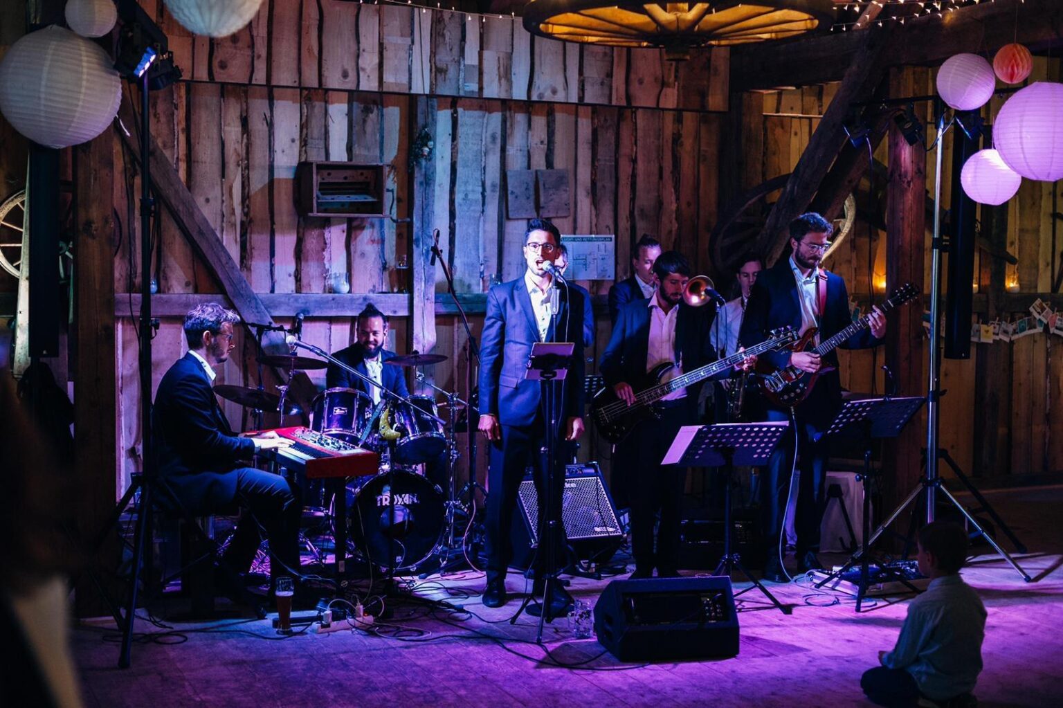 Die Brass Tacks Band spielt live bei einem traditionellen Stadl-Event, das Publikum feiert ausgelassen in rustikalem Ambiente.