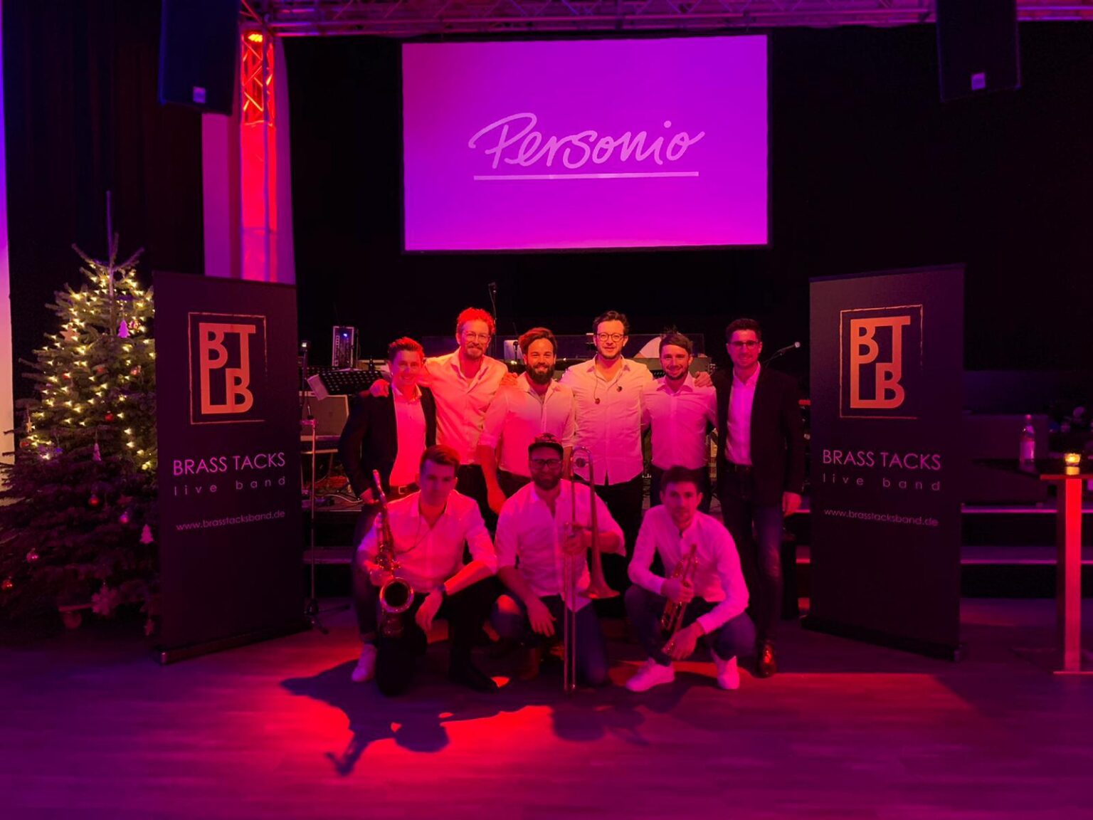 Die Brass Tacks Band spielt live auf einer festlich dekorierten Bühne bei einer Weihnachtsfeier einer Firma, umgeben von Weihnachtslichtern und fröhlichen Gästen.