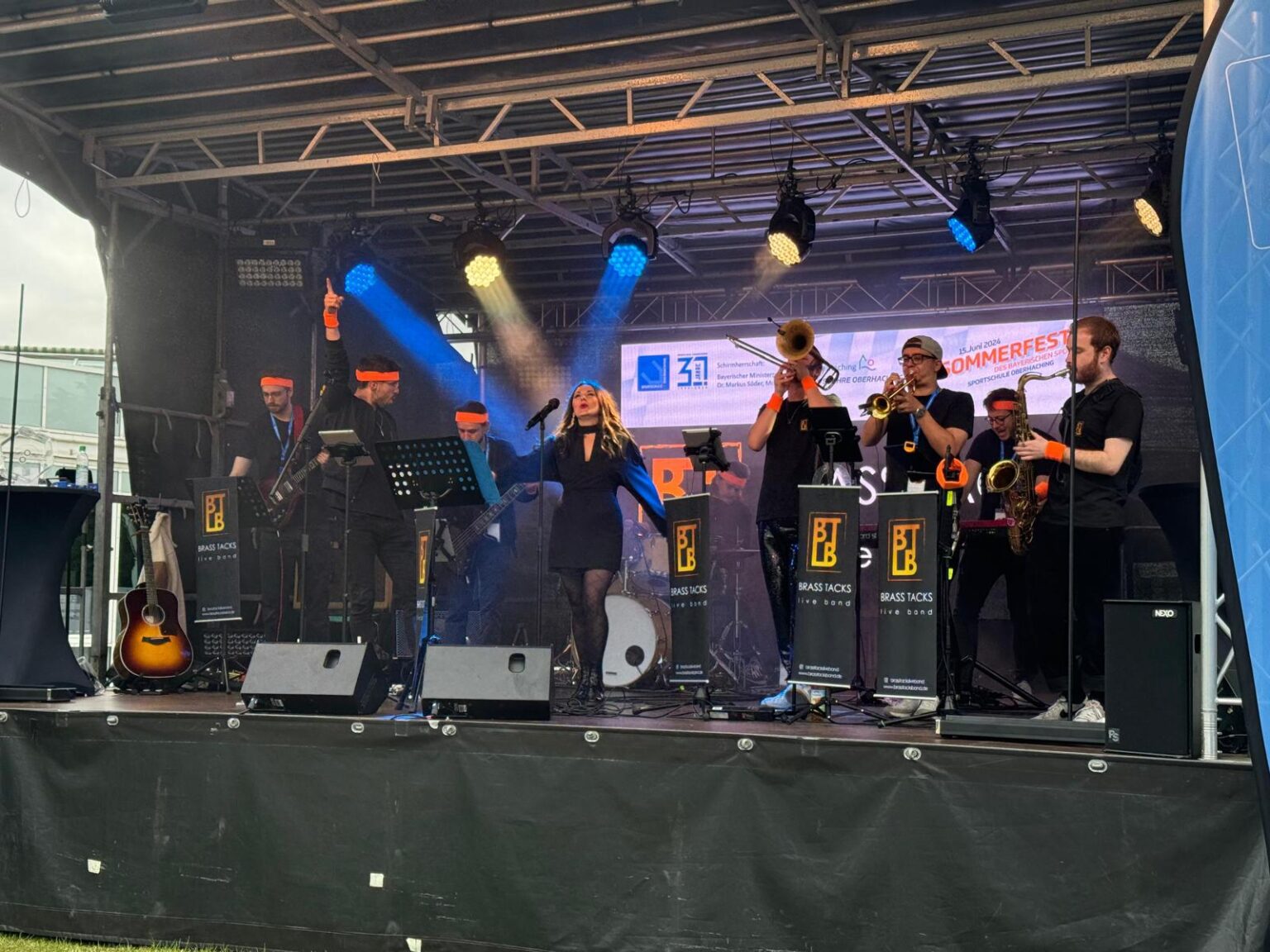 Die Brass Tacks Band spielt live bei einem Sportfest, das Publikum feiert ausgelassen und die Stimmung ist voller Energie und Freude.