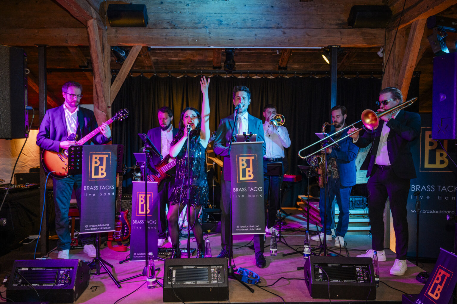 Die komplette Brass Tacks Band – mit männlichem und weiblichem Lead-Gesang – performt stilvoll und energiegeladen auf einer Bühne bei einem festlichen Gala-Event.